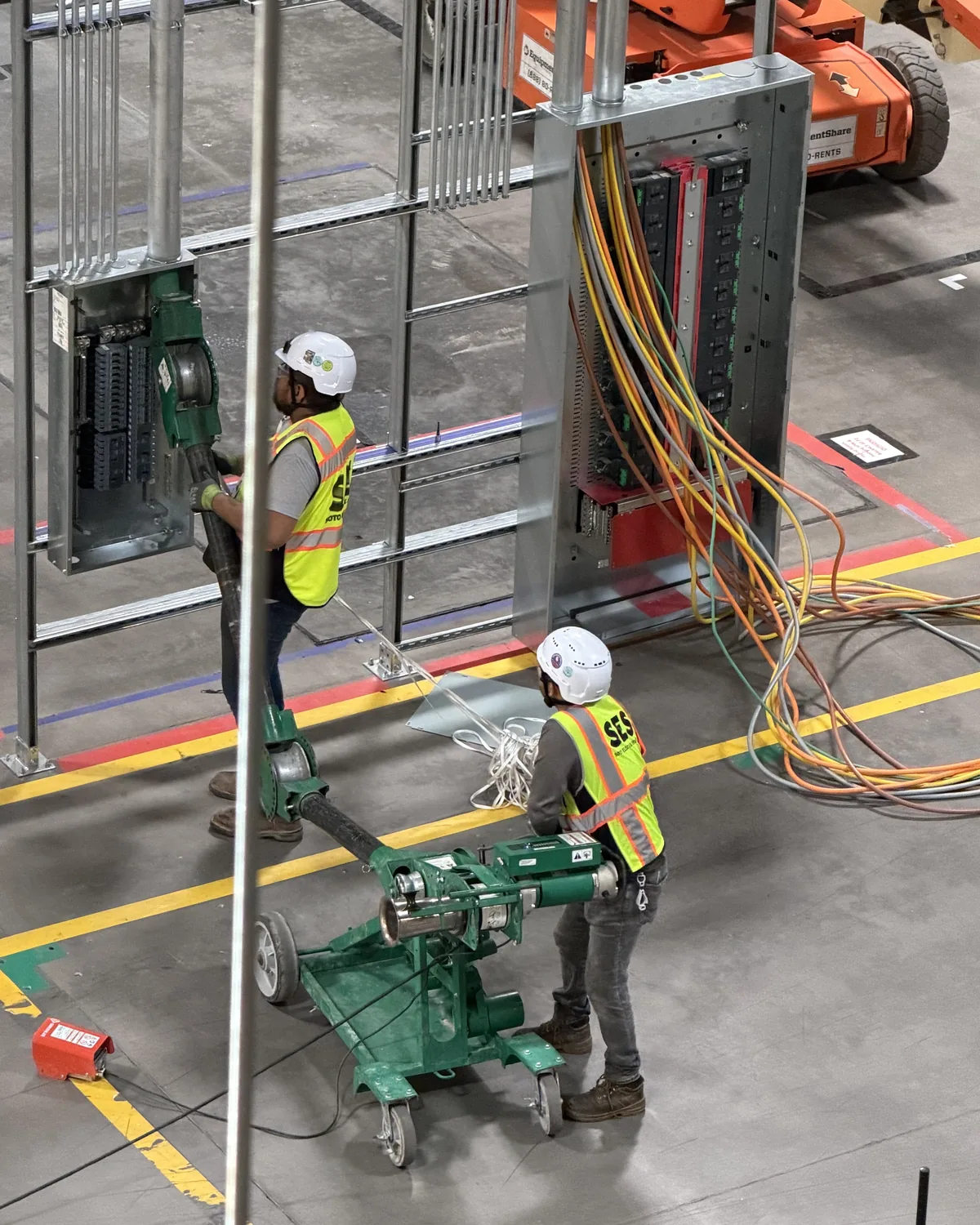 Two electricians in hard hats and high-visibility vests working together with a material lift beneath overhead cable tray